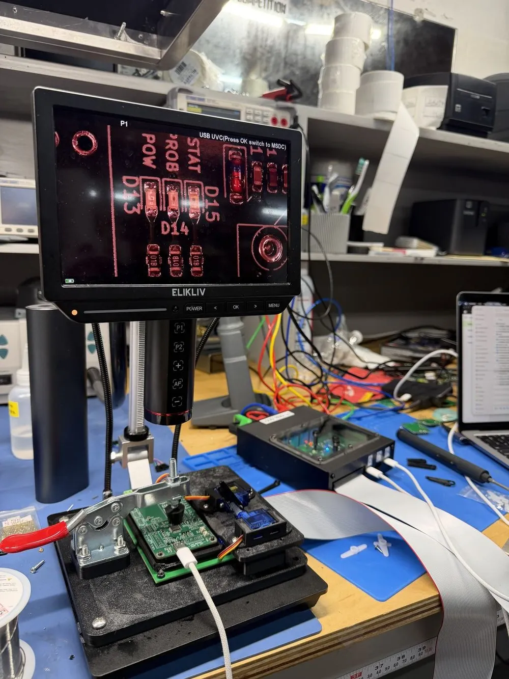 Close view of the camera head mounted over a test fixture, with a board clamped below it and a monitor above showing the camera’s view.